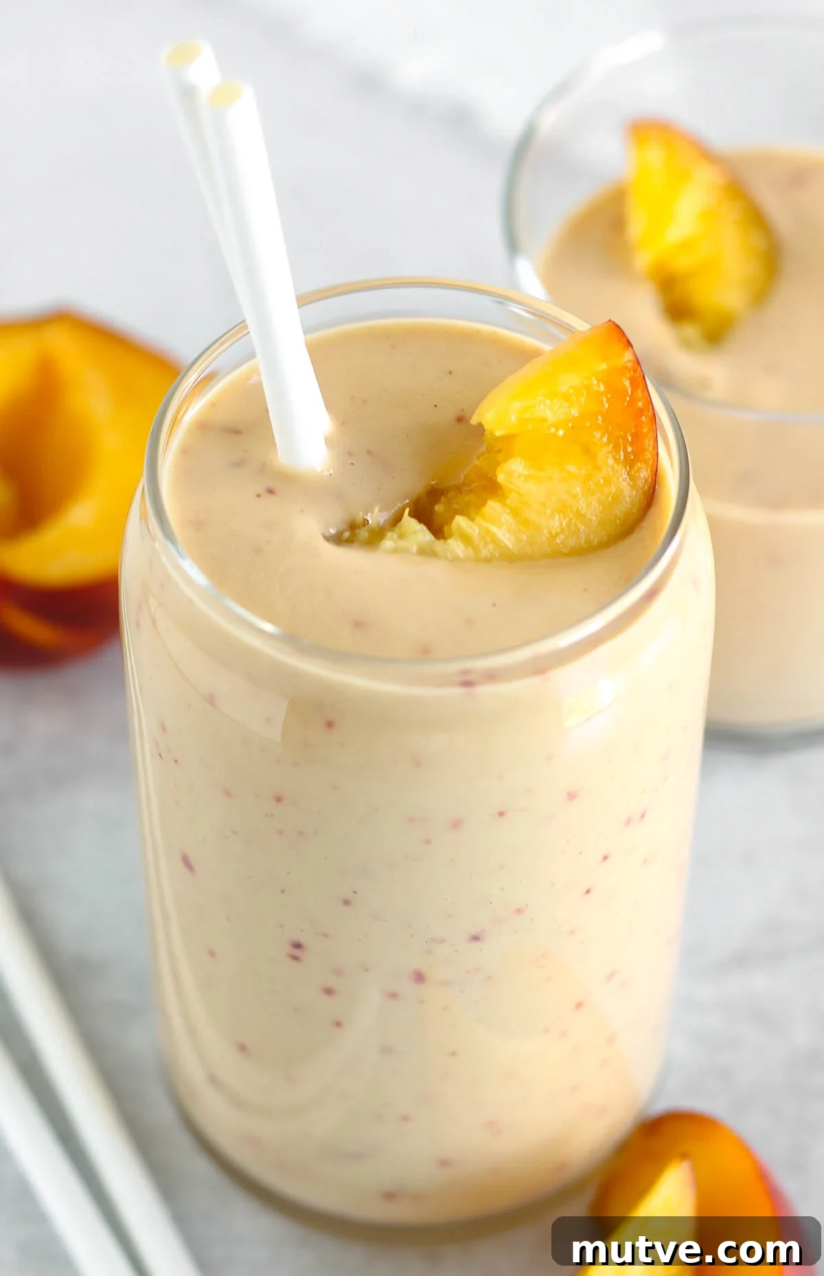 Tall glass filled with a peach banana smoothie topped with a peach slice and two straws.