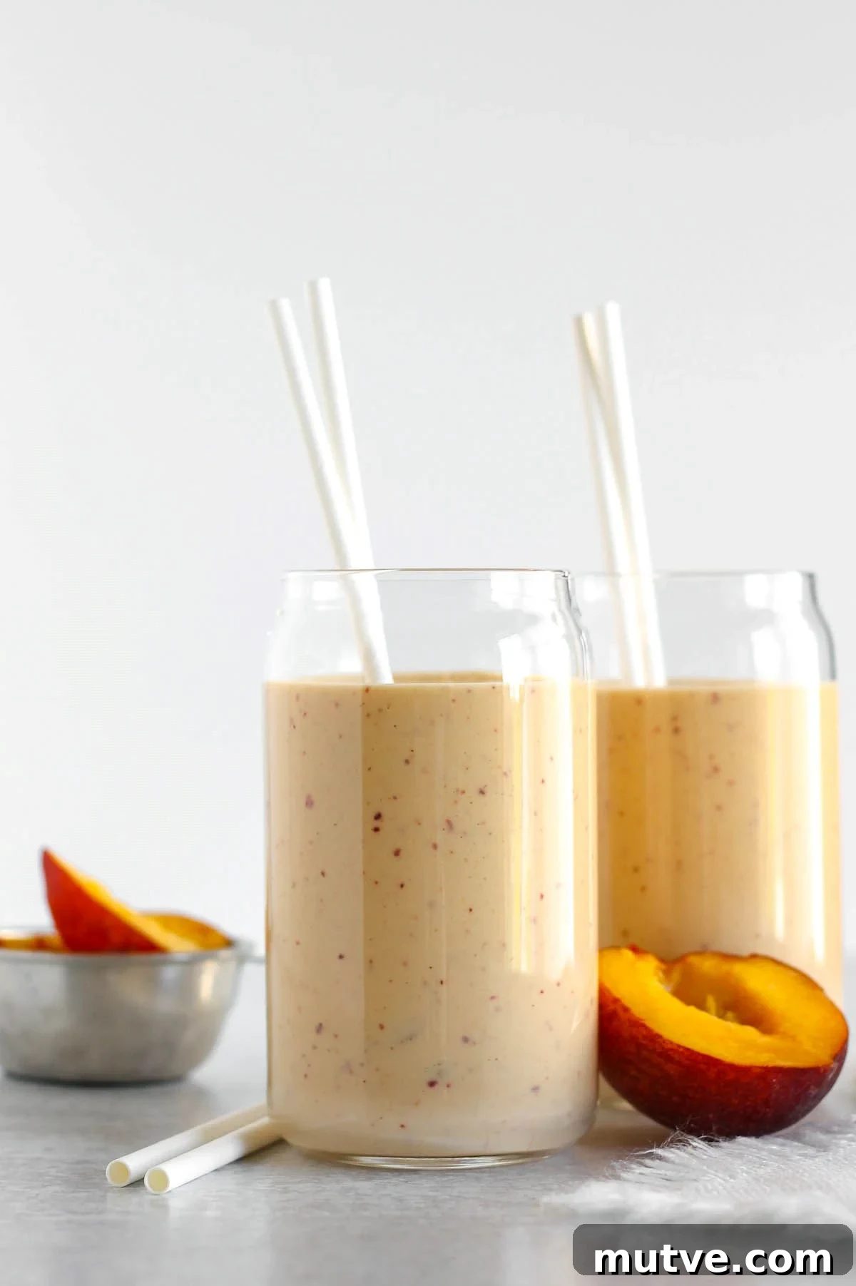 Two glasses filled with peach banana smoothie with two straws in each glass and half a peach laying next to the glasses.
