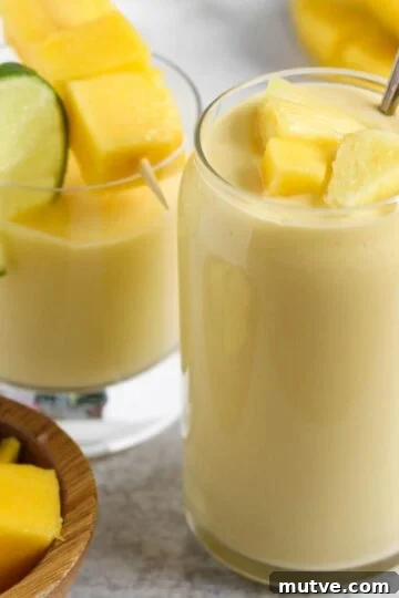 Two glasses filled with a creamy yellow smoothie topped with fresh pineapple and mangos.