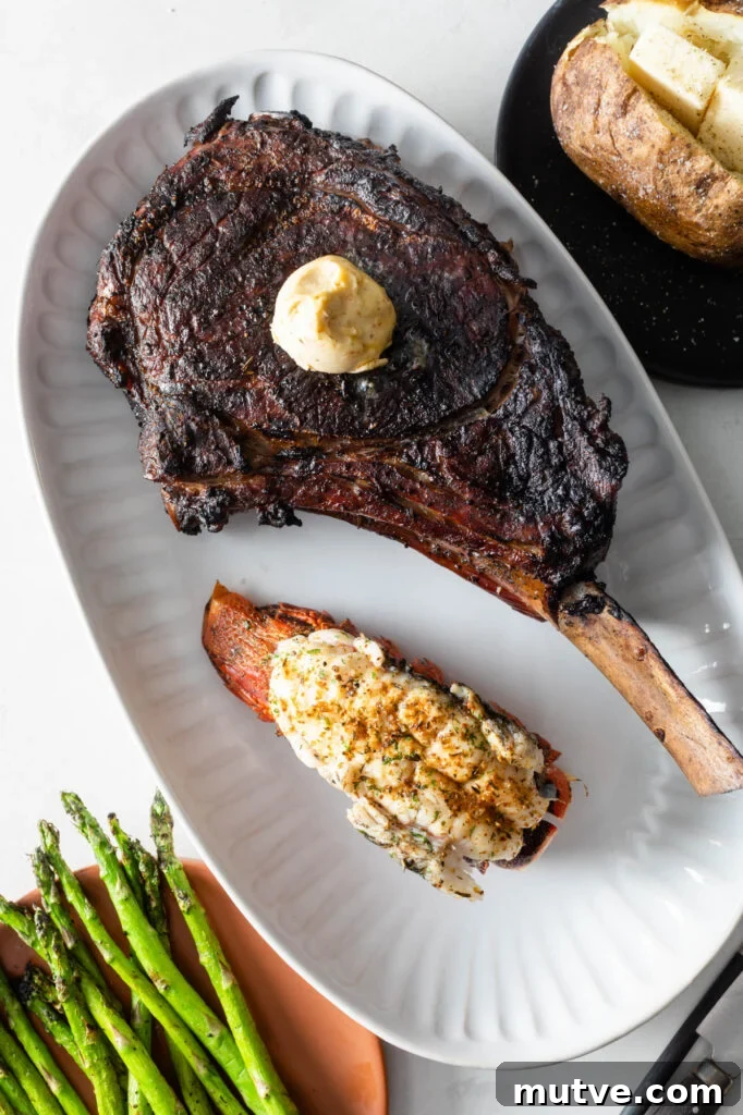 cooked tomahawk steak topped with a scoop of compound butter on a plate with a cooked lobster tail. asparagus spears and a baked potato with butter in the background