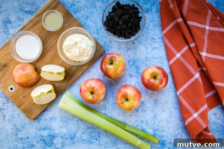 Overhead view of 4 5 apples (one chopped), two stalks of celery, a bowl of raisins, a bowl with mayonnaise, a small bowl with lemon juice, and a small bowl with sugars all on a blue surface with a coral plaid towel to the side