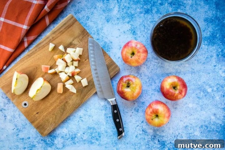 Overhead view of a chopping board with shopped apple and a knife, four apples to the side, a bowl of raisins soaking in water, all on a blue surface with a coral towel laying to the side.